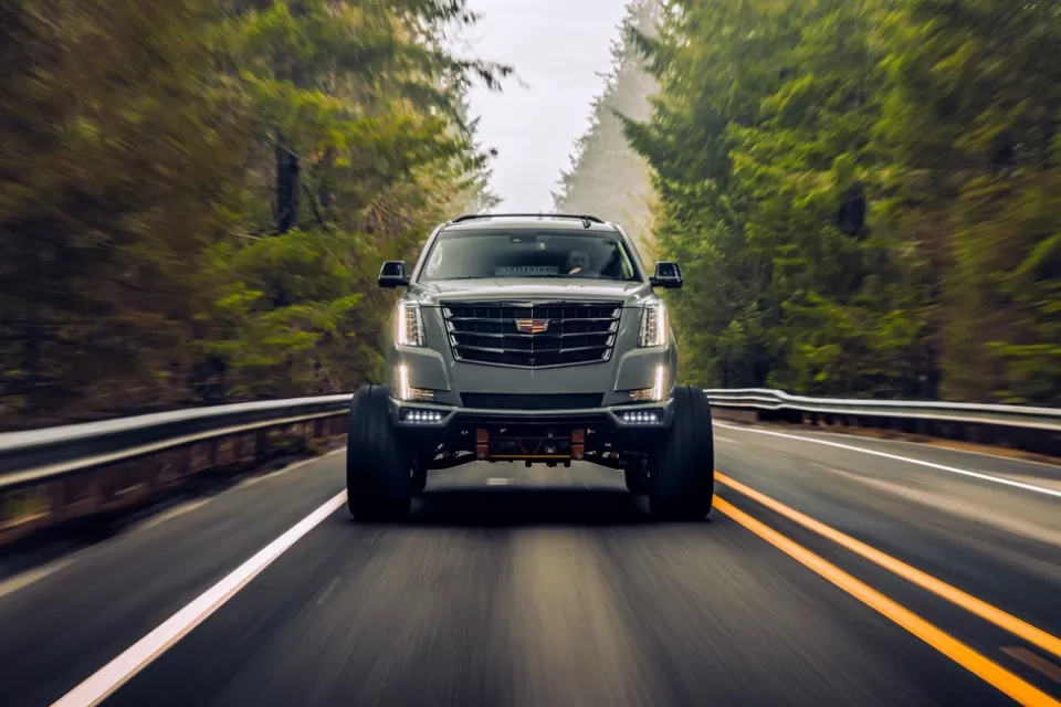 Cadillac Escalade SUV driving toward camera on black asphalt road surrounded by forest